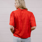 A woman viewed from behind wearing a bright red short-sleeve blouse with gathered detailing and light-wash jeans, standing against a white paneled wall.