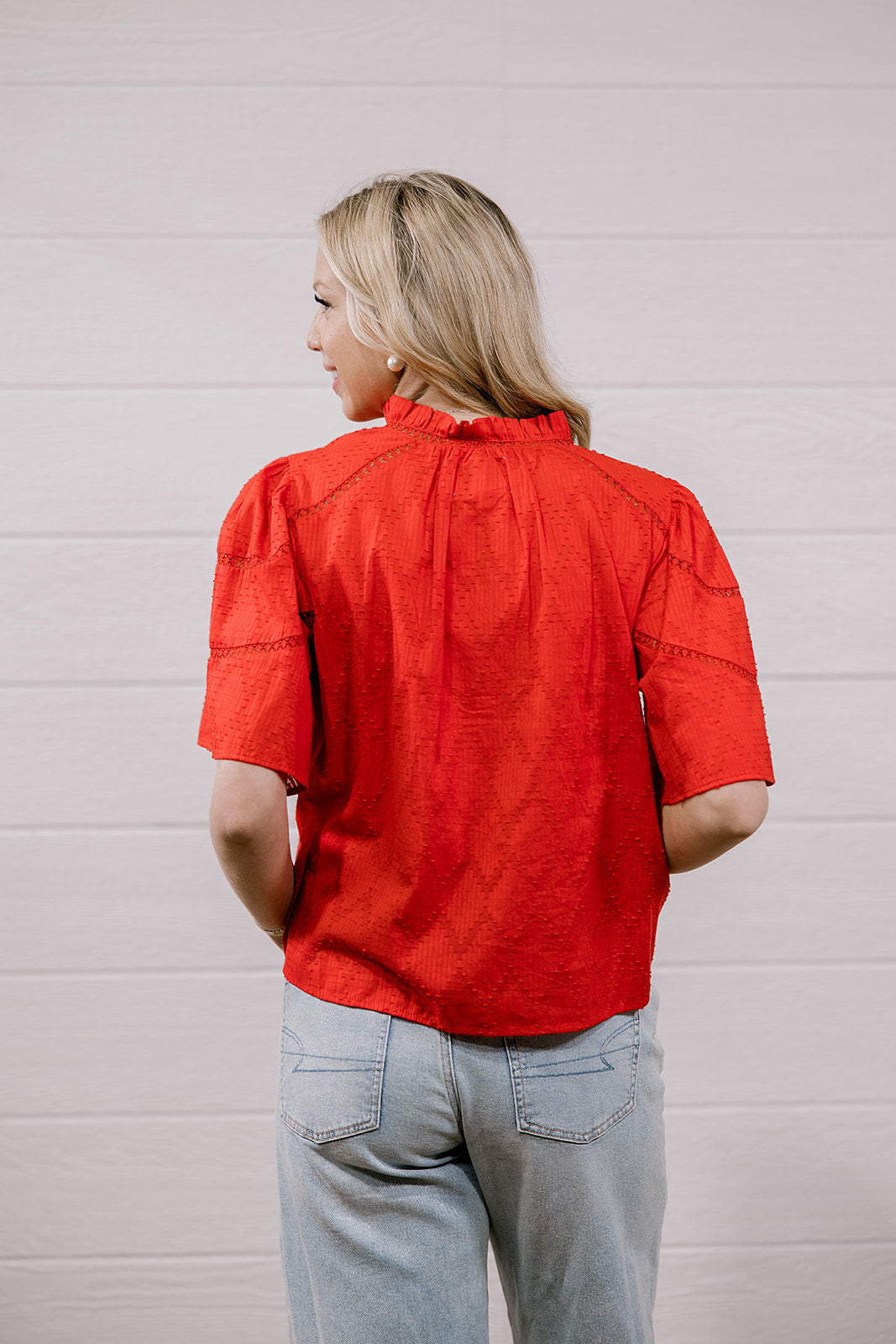 A woman viewed from behind wearing a bright red short-sleeve blouse with gathered detailing and light-wash jeans, standing against a white paneled wall.