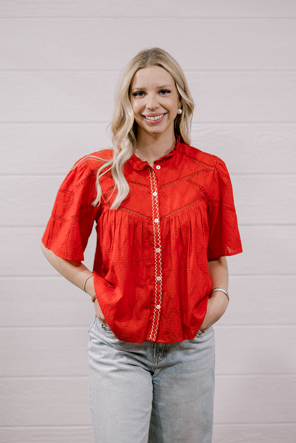 A woman with blonde hair wearing a bright red short-sleeve embroidered blouse and light-wash jeans, standing against a white paneled wall.