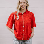 A woman with blonde hair wearing a bright red short-sleeve embroidered blouse and light-wash jeans, standing against a white paneled wall.
