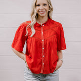 A woman with blonde hair wearing a bright red short-sleeve embroidered blouse and light-wash jeans, standing against a white paneled wall.