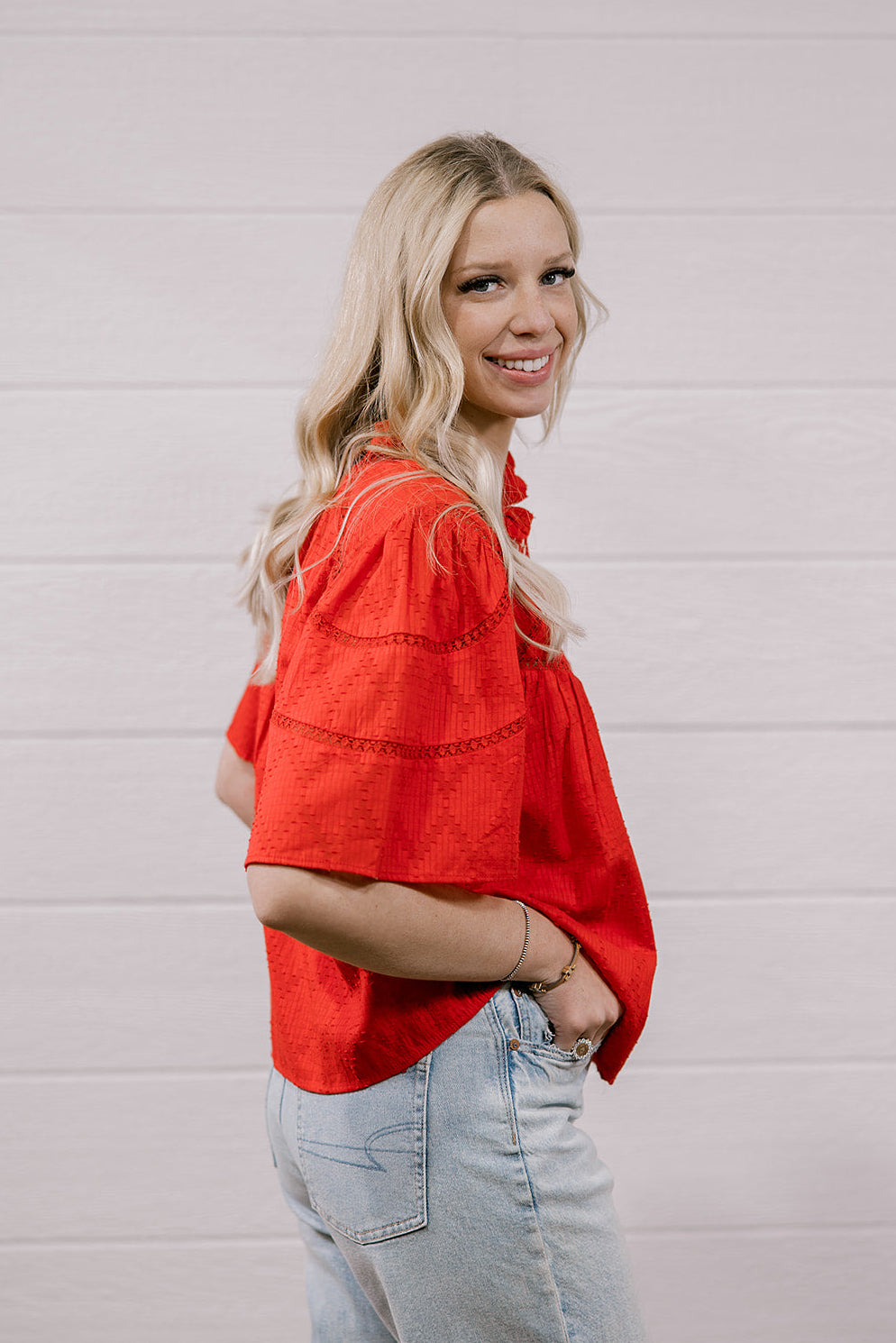 A woman with blonde hair wearing a bright red short-sleeve blouse and light blue jeans, standing against a white paneled wall.