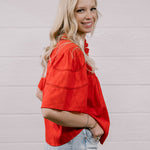 A woman with blonde hair wearing a bright red short-sleeve blouse and light blue jeans, standing against a white paneled wall.