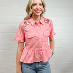 Women wearing a pink gingham pearl snap top with buckin cowgirl girl embroidered on shoulders and ruffles at sleeves and waist. 