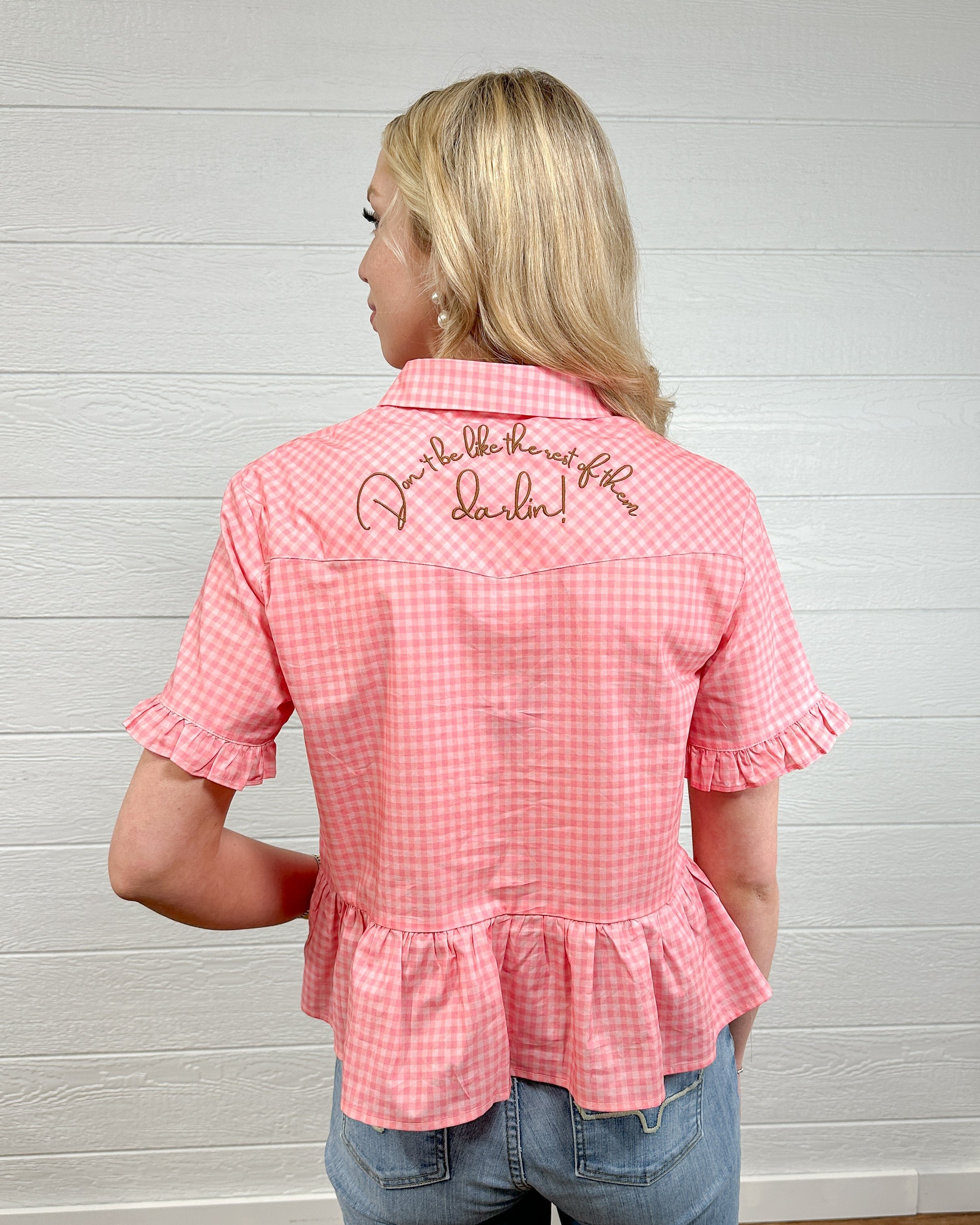 Back view women wearing a pink gingham pearl snap top with embroidered on back and ruffles at sleeves and waist. 
