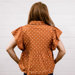 A woman viewed from behind wearing a rust-orange short-sleeve blouse with white polka dots and a gathered peplum waist, paired with dark blue jeans, standing against a white paneled wall.