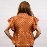 A woman viewed from behind wearing a rust-orange short-sleeve blouse with white polka dots and a gathered peplum waist, paired with dark blue jeans, standing against a white paneled wall.