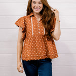 A smiling young woman with long brown hair stands against a white shiplap wall, wearing a rust-orange short-sleeve blouse with white polka dots and white vertical trim, paired with dark blue jeans.
