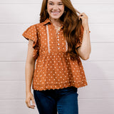 A smiling young woman with long brown hair stands against a white shiplap wall, wearing a rust-orange short-sleeve blouse with white polka dots and white vertical trim, paired with dark blue jeans.