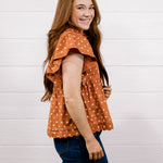 A woman with long light brown hair is shown from the side/back wearing a rust-orange short-sleeve blouse with white polka dots and dark blue jeans, standing against a white wood-paneled wall.