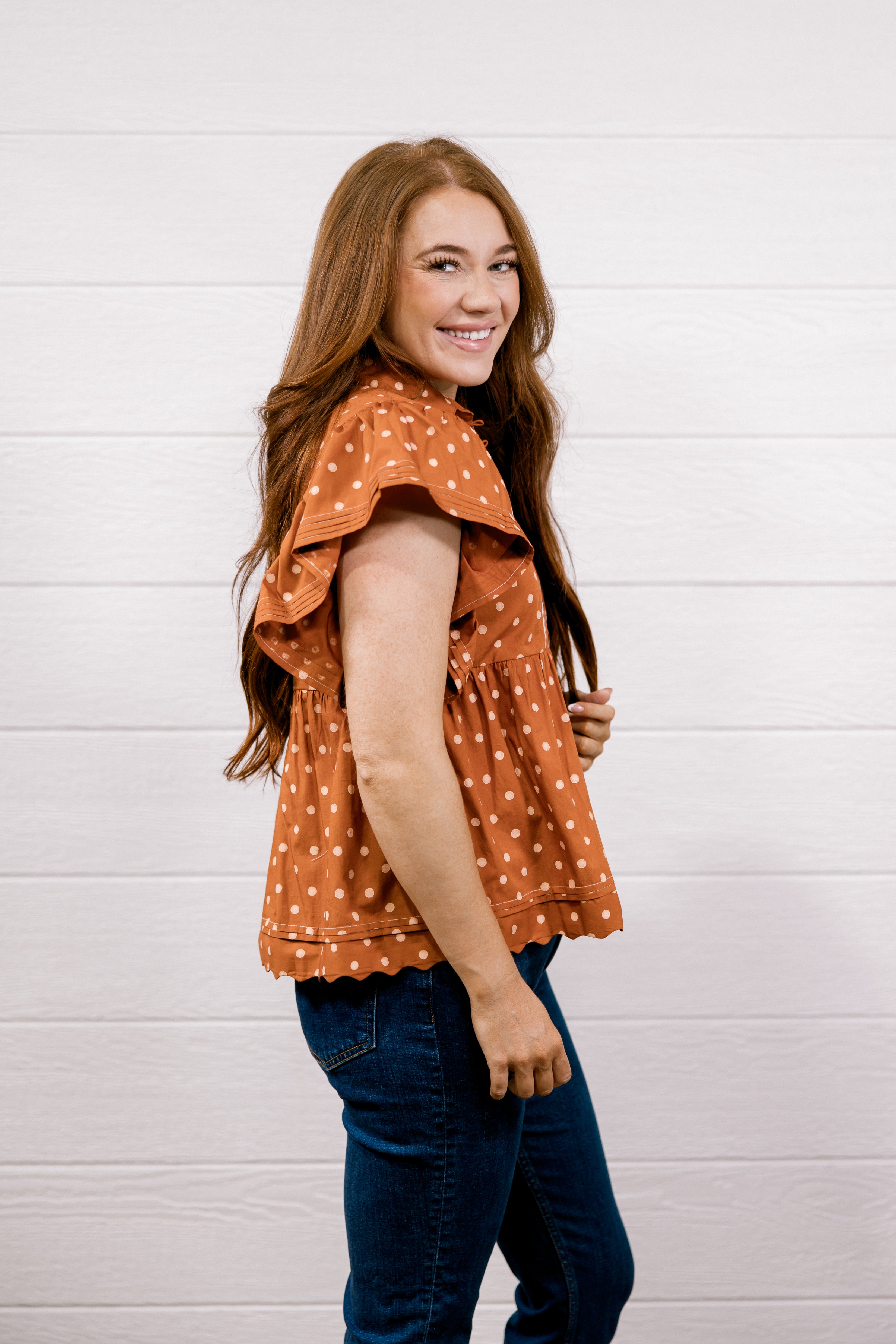 A woman with long light brown hair is shown from the side/back wearing a rust-orange short-sleeve blouse with white polka dots and dark blue jeans, standing against a white wood-paneled wall.