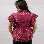 A woman viewed from behind wearing a maroon short-sleeve blouse with white polka dots and dark blue jeans, standing against a white paneled wall.