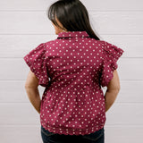 A woman viewed from behind wearing a maroon short-sleeve blouse with white polka dots and dark blue jeans, standing against a white paneled wall.