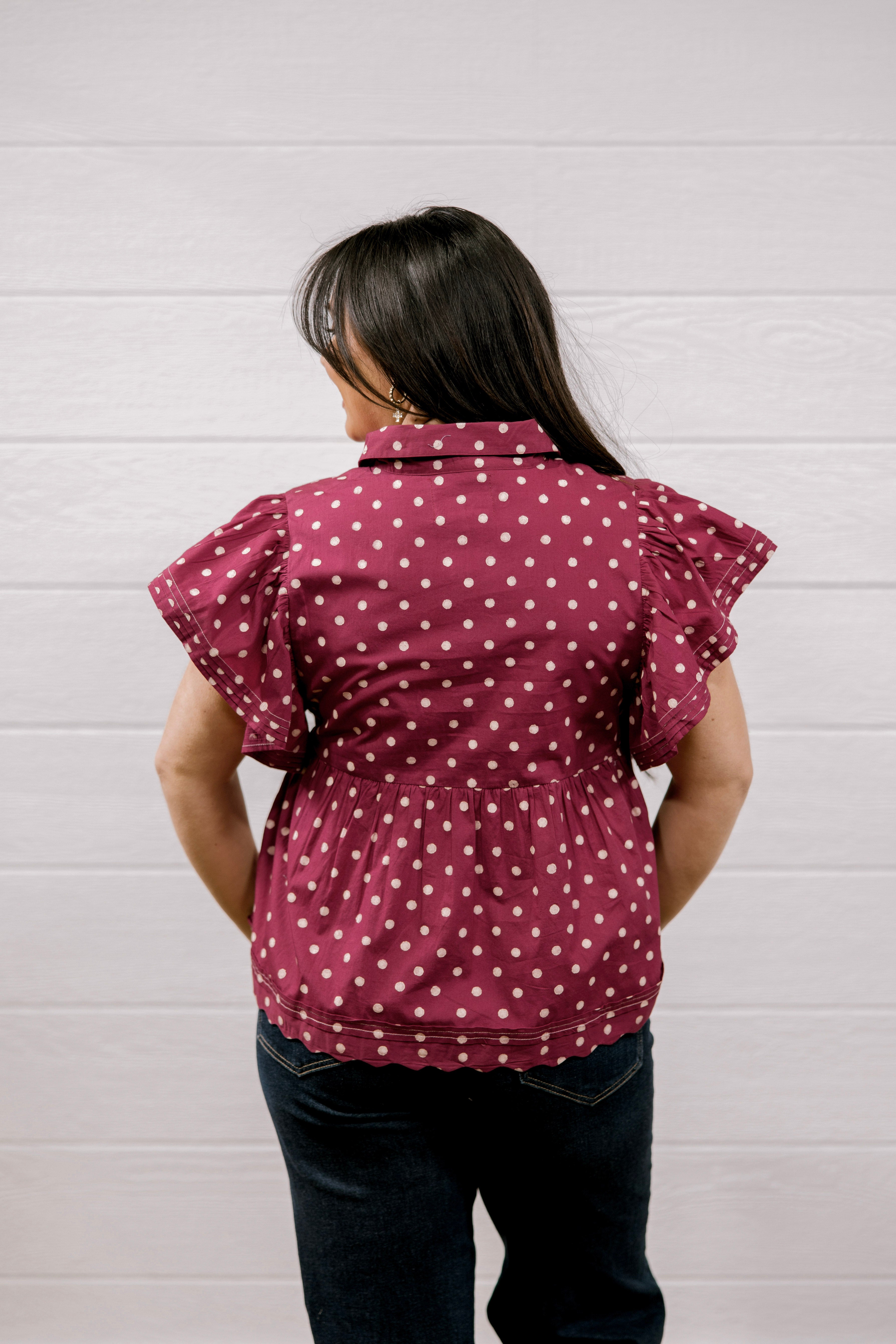A woman viewed from behind wearing a maroon short-sleeve blouse with white polka dots and dark blue jeans, standing against a white paneled wall.