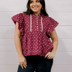 A woman modeling a maroon short-sleeve blouse with white polka dots and white embroidered details on the front, paired with dark blue jeans, standing against a white wood-paneled wall.