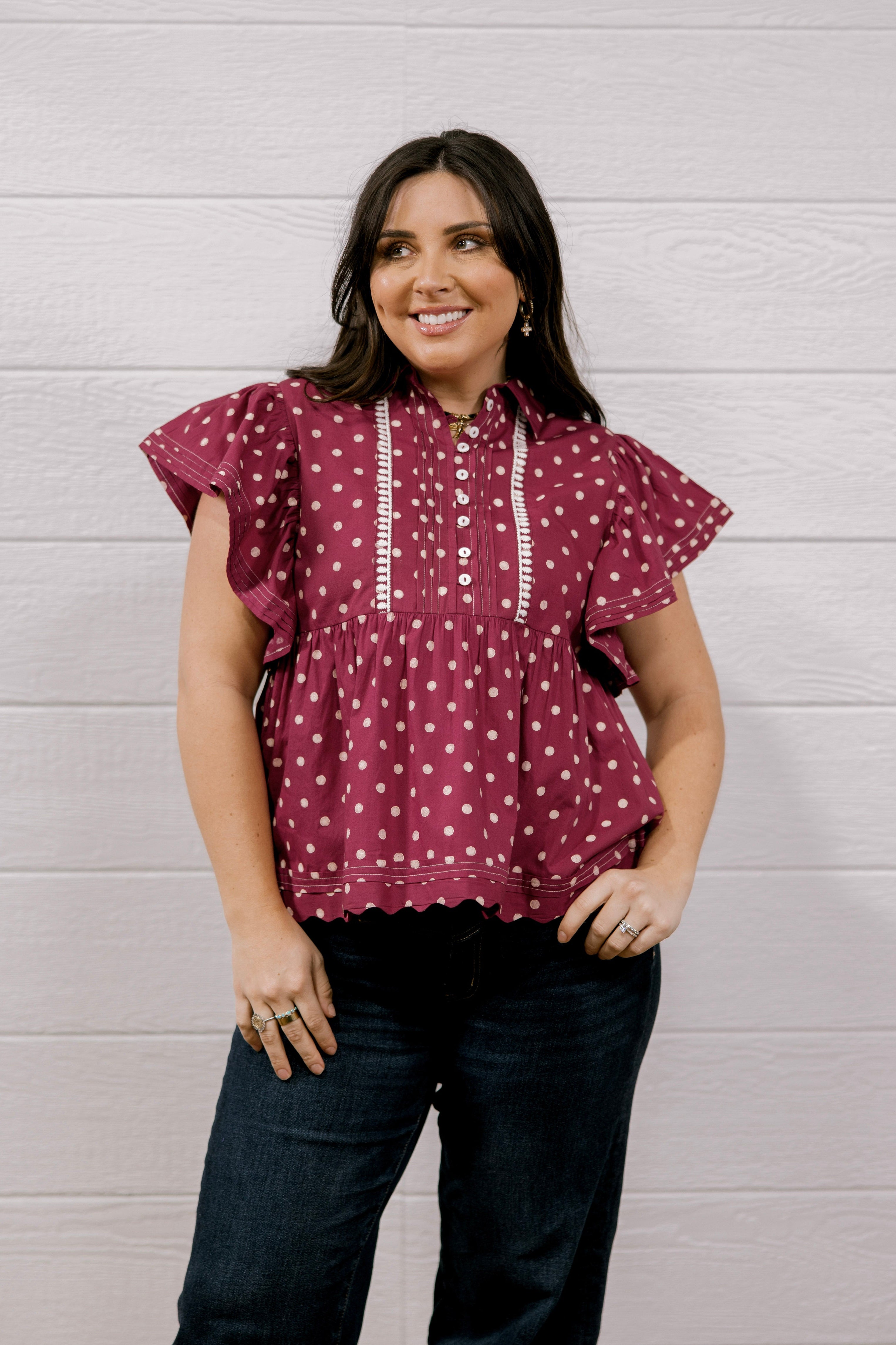 A woman modeling a maroon short-sleeve blouse with white polka dots and white embroidered details on the front, paired with dark blue jeans, standing against a white wood-paneled wall.