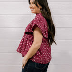 A woman with long dark hair is shown from the side, wearing a short-sleeve ruffled blouse in maroon with white polka dots and dark blue jeans, standing against a white wood-paneled wall.