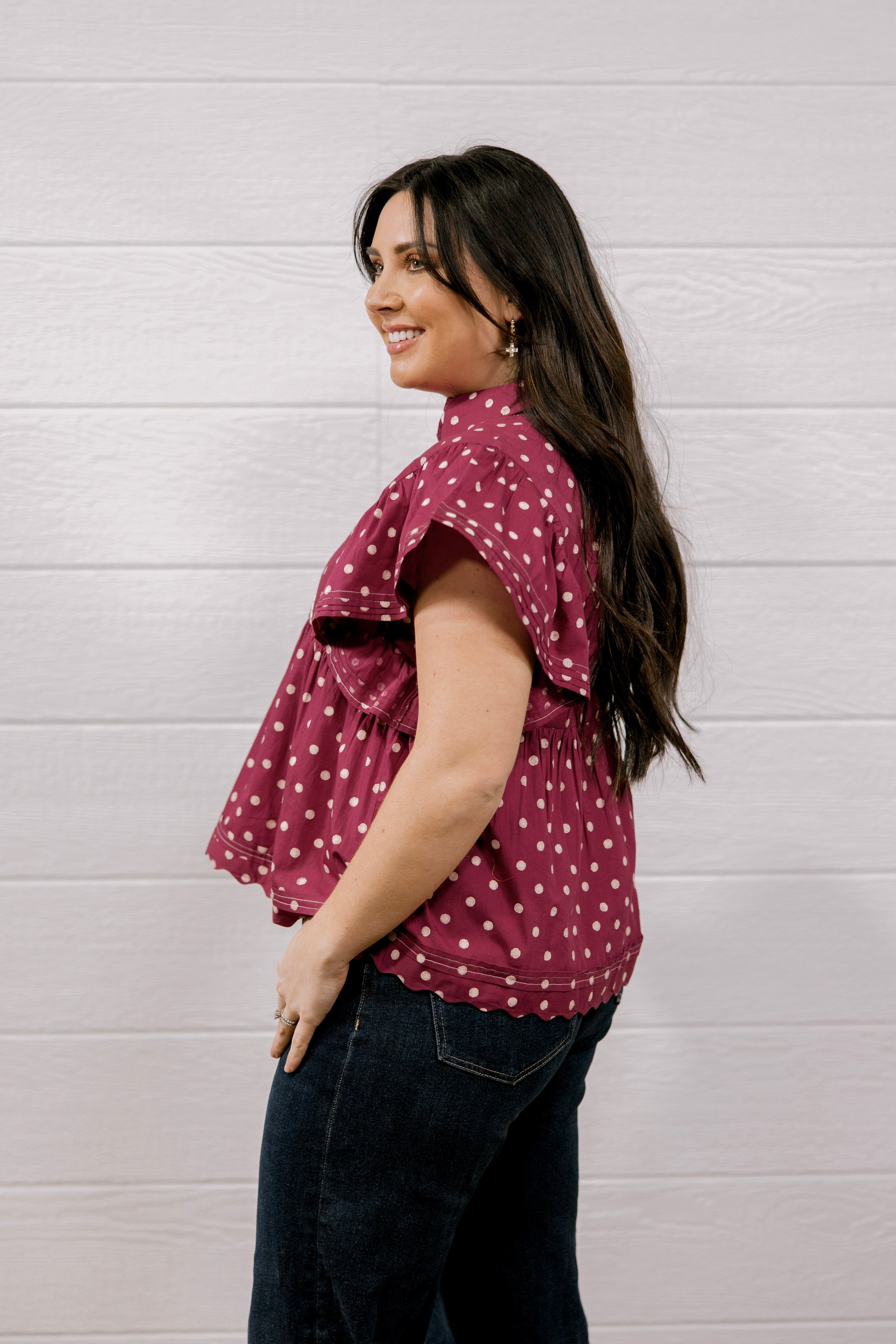 A woman with long dark hair is shown from the side, wearing a short-sleeve ruffled blouse in maroon with white polka dots and dark blue jeans, standing against a white wood-paneled wall.