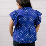 A woman viewed from the back wearing a purple short-sleeve blouse with white polka dots and ruffled sleeves, paired with dark jeans, standing against a white paneled wall.
