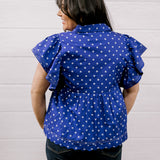 A woman viewed from the back wearing a purple short-sleeve blouse with white polka dots and ruffled sleeves, paired with dark jeans, standing against a white paneled wall.