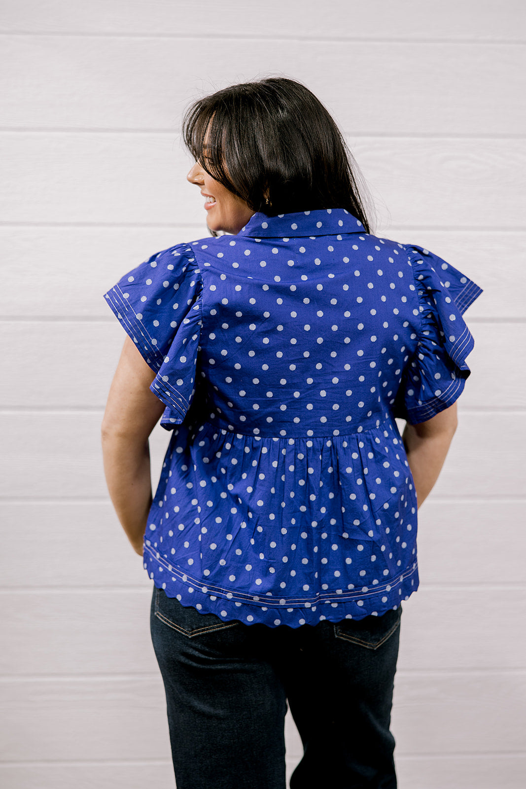 A woman viewed from the back wearing a purple short-sleeve blouse with white polka dots and ruffled sleeves, paired with dark jeans, standing against a white paneled wall.