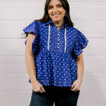 A woman wearing a purple short-sleeve blouse with white polka dots and white embroidered trim, paired with dark jeans, standing against a white wood-paneled wall.