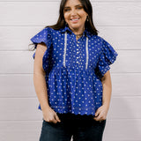 A woman wearing a purple short-sleeve blouse with white polka dots and white embroidered trim, paired with dark jeans, standing against a white wood-paneled wall.