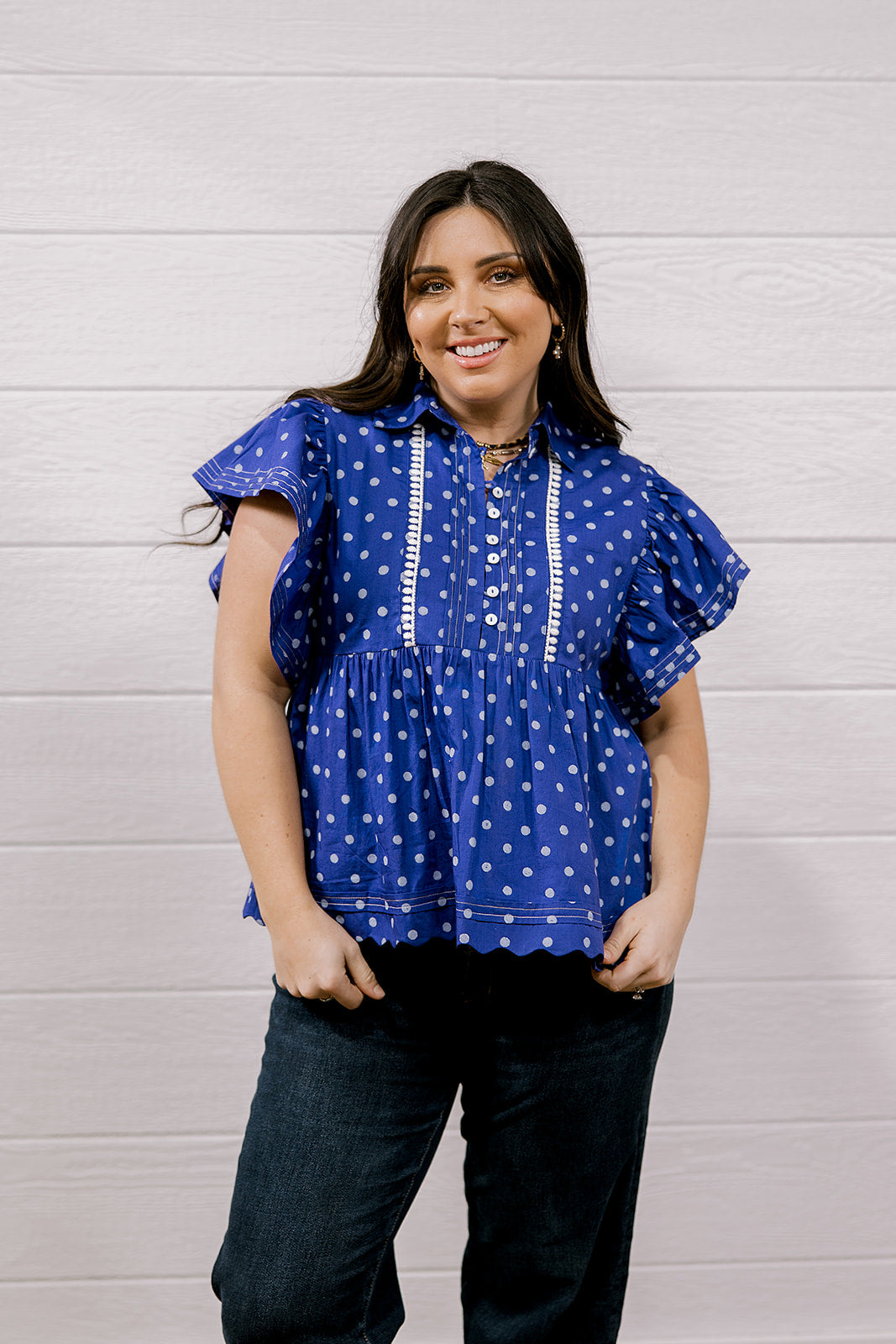 A woman wearing a purple short-sleeve blouse with white polka dots and white embroidered trim, paired with dark jeans, standing against a white wood-paneled wall.