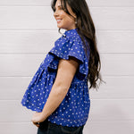 A woman wearing a purple short-sleeve top with white polka dots and dark jeans, standing against a white paneled wall.