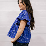 A woman wearing a purple short-sleeve top with white polka dots and dark jeans, standing against a white paneled wall.