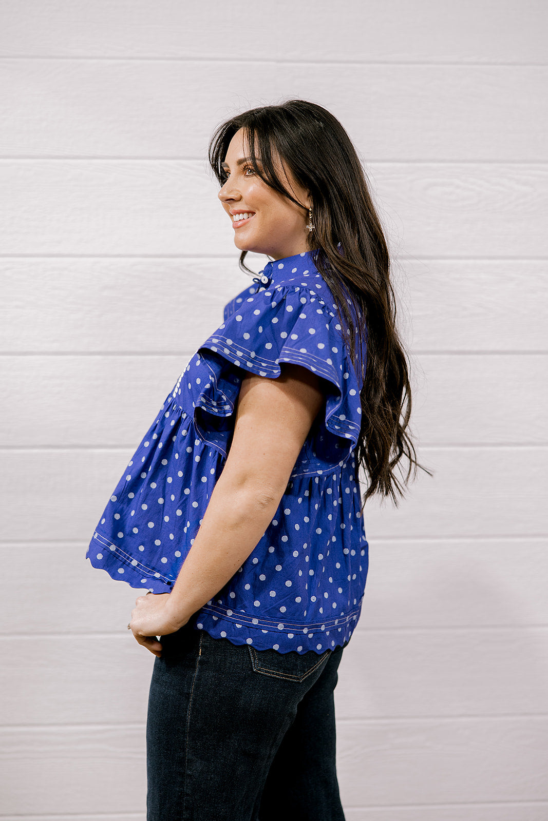 A woman wearing a purple short-sleeve top with white polka dots and dark jeans, standing against a white paneled wall.