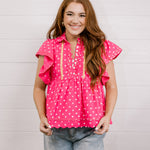 Woman wearing a pink polka dot blouse with ruffled sleeves against a light background facing front. 