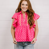 Woman wearing a pink polka dot blouse with ruffled sleeves against a light background facing front. 