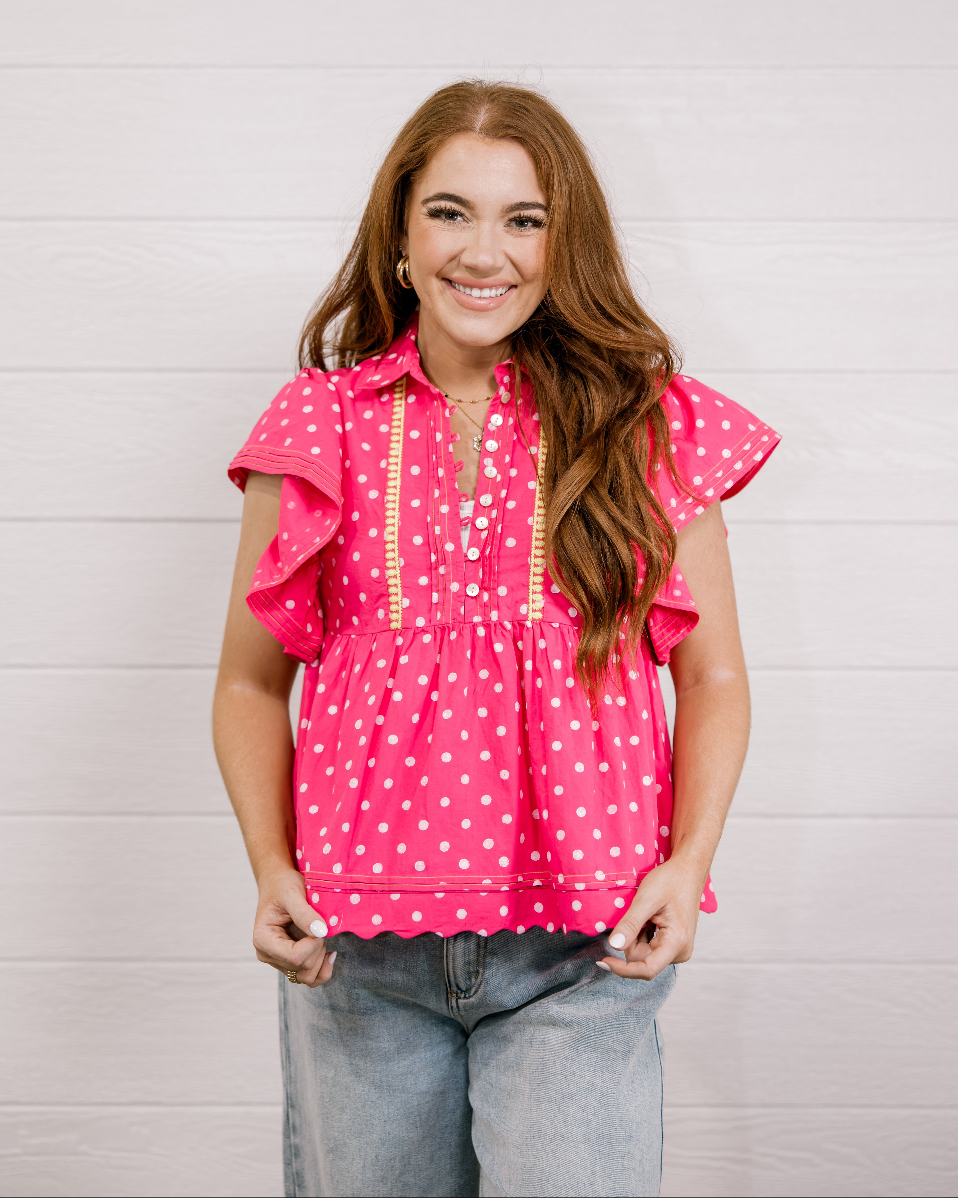 Woman wearing a pink polka dot blouse with ruffled sleeves against a light background facing front. 