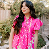 Pink top with white polk a dots and yellow accent stitching on women in a garden. 