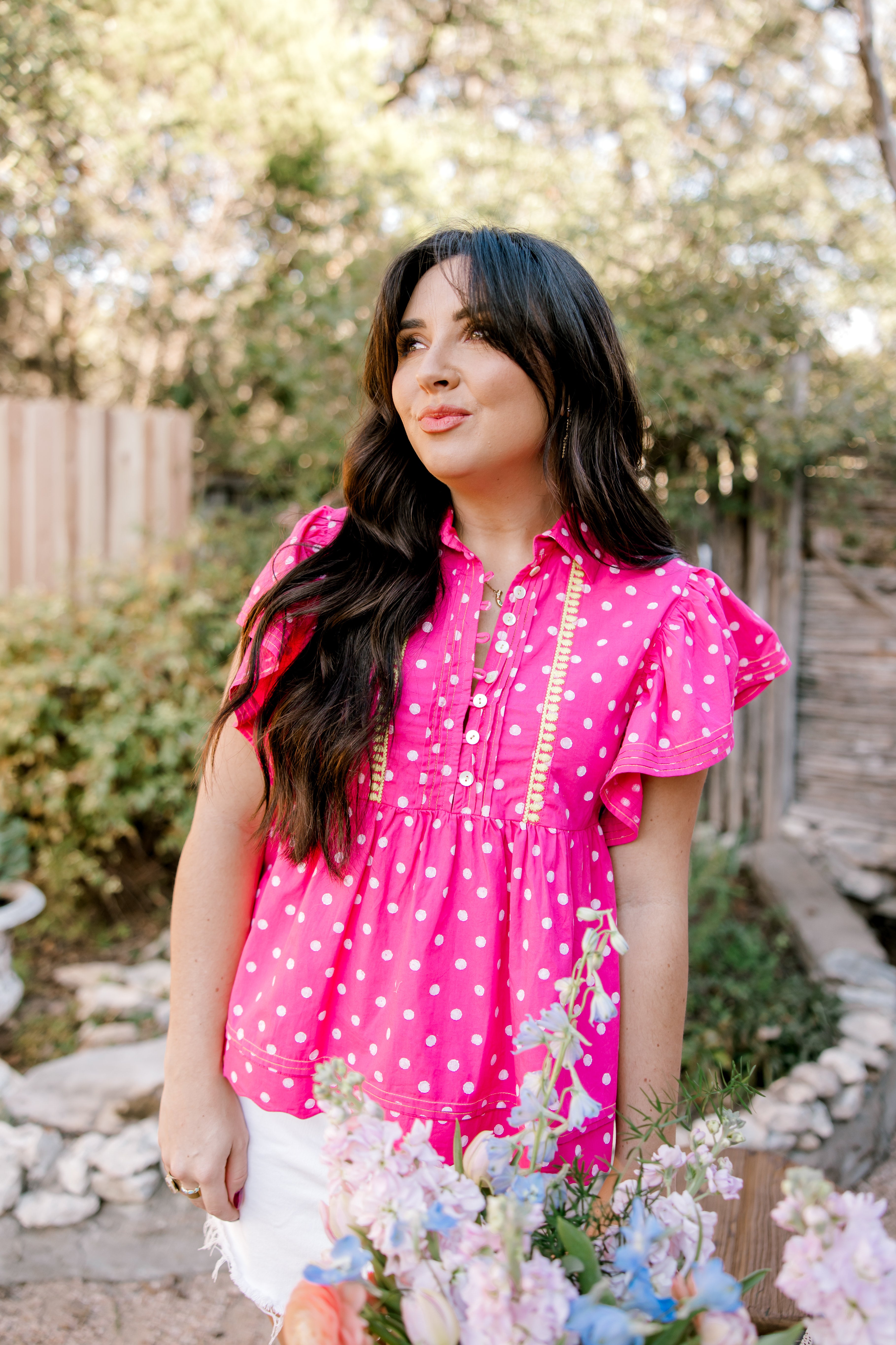 Pink top with white polk a dots and yellow accent stitching on women in a garden. 
