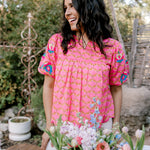 Woman in a pink dress with floral patterns standing behind a table with flowers outdoors.