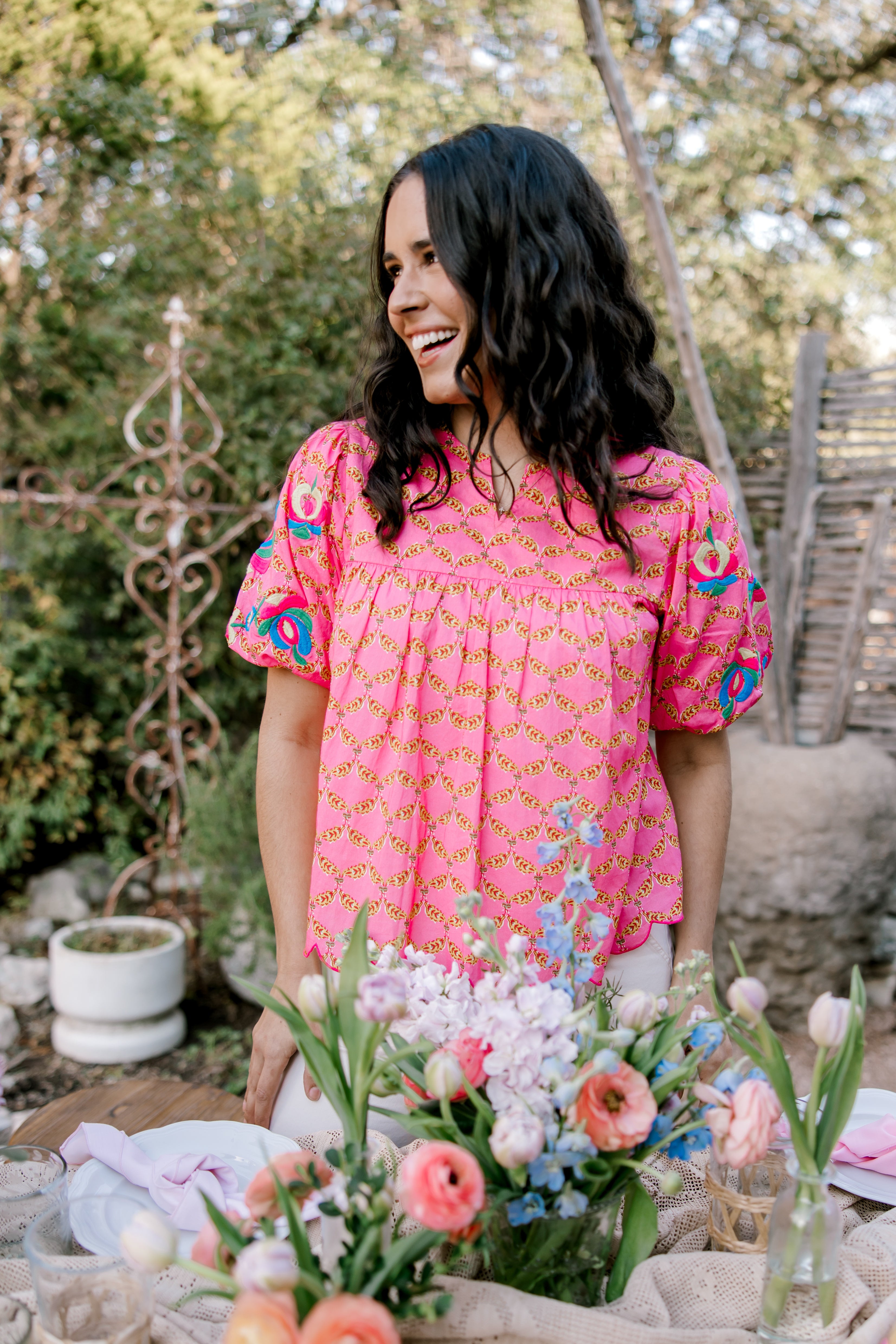 Woman in a pink dress with floral patterns standing behind a table with flowers outdoors.