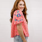 Woman wearing a pink embroidered top against a white background facing the side. 