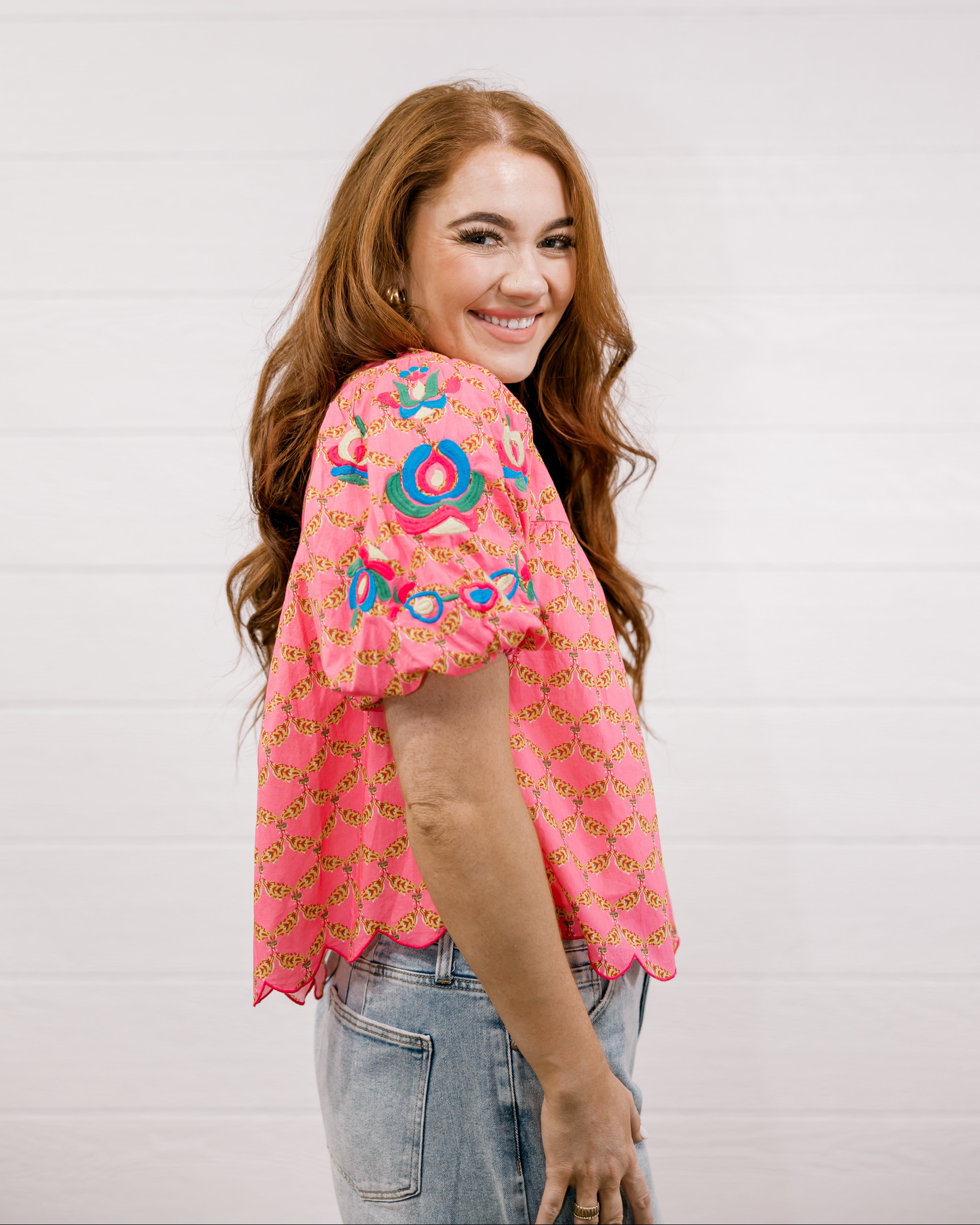 Woman wearing a pink embroidered top against a white background facing the side. 