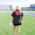 Woman with blonde hair standing in a back profile position wearing the Reece Top in a lifestyle scene. The focal point of this image is the red floral embroidery on back of top as well as pleating on back. The model paired this top with black denim shorts. 