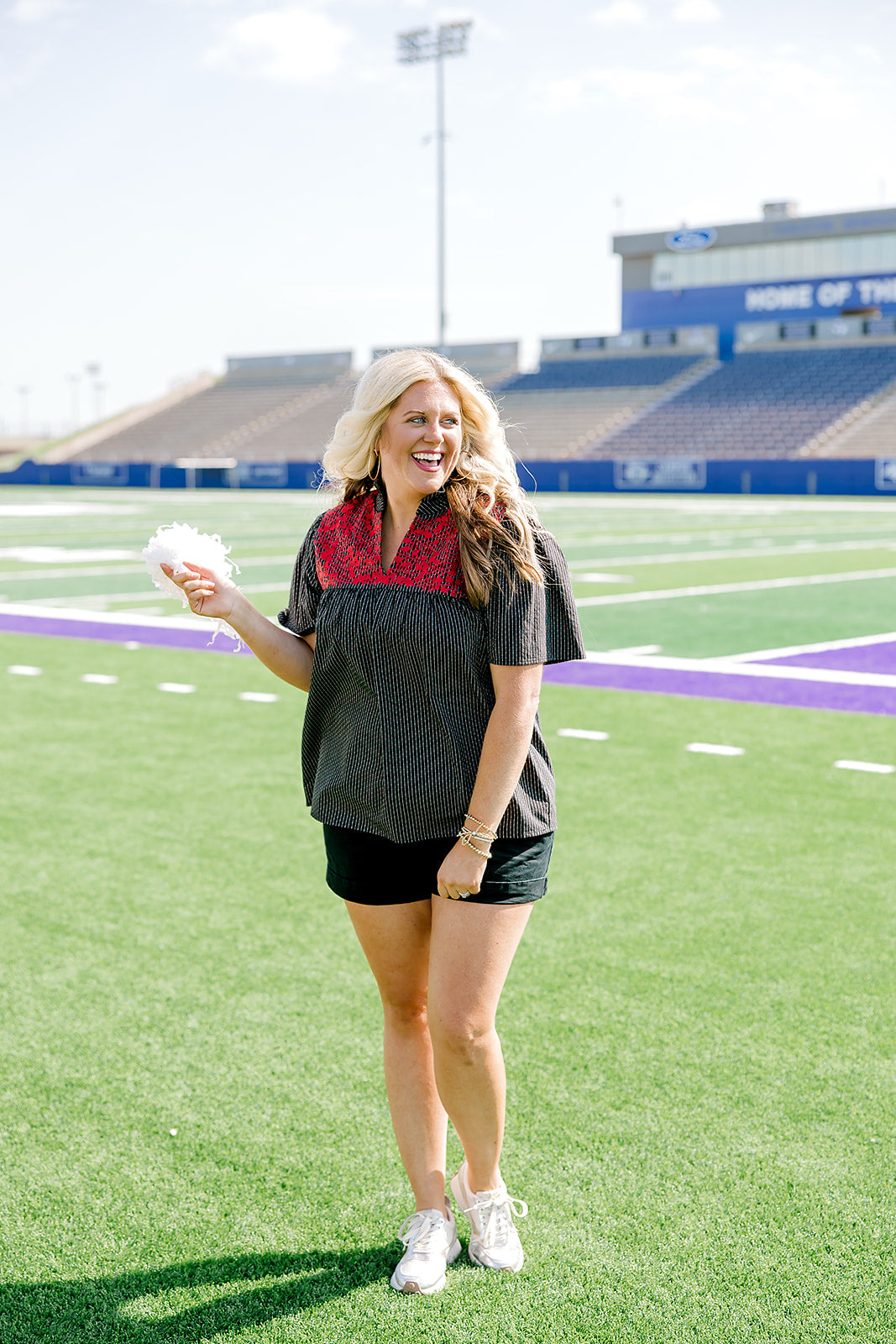 Woman with blonde hair standing in a front profile position wearing the Reece Top in a lifestyle scene. The focal point of this image is the red floral embroidery on front of top as well as v-neckline. The model paired this top with black denim shorts. 