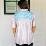 Woman with dark hair wearing a top in the Reece body style. Fabric is pink iridescent and is accented with turquoise embroidery along shoulders and chest. Model paired this top with faux leather leggings. 