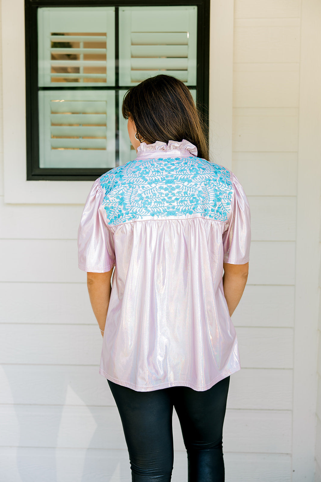 Woman with dark hair wearing a top in the Reece body style. Fabric is pink iridescent and is accented with turquoise embroidery along shoulders and chest. Model paired this top with faux leather leggings. 