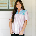 Woman with dark hair wearing a top in the Reece body style. Fabric is pink iridescent and is accented with turquoise embroidery along shoulders and chest. Model paired this top with faux leather leggings. 