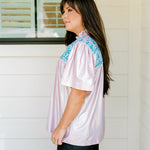 Woman with dark hair wearing a top in the Reece body style. Fabric is pink iridescent and is accented with turquoise embroidery along shoulders and chest. Model paired this top with faux leather leggings. 