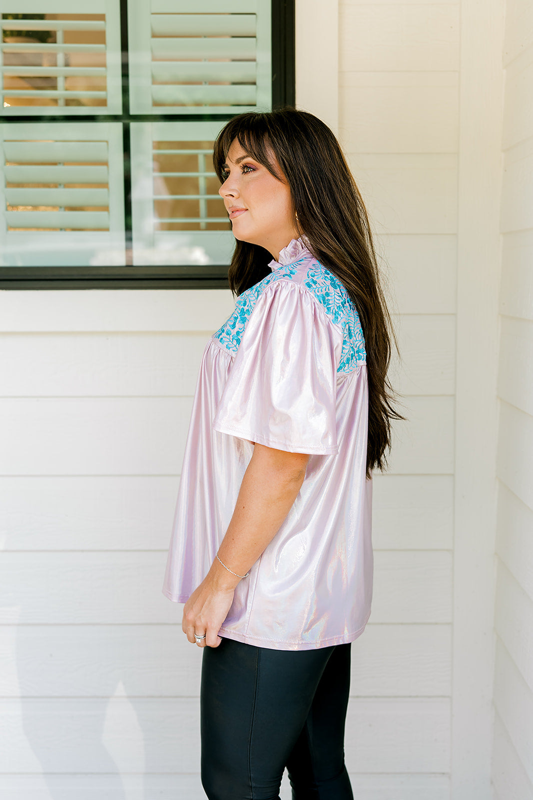 Woman with dark hair wearing a top in the Reece body style. Fabric is pink iridescent and is accented with turquoise embroidery along shoulders and chest. Model paired this top with faux leather leggings. 