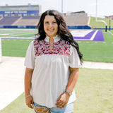 Woman with dark hair standing in a front profile position wearing the Reece Top in a lifestyle scene. The focal point of this image is the ruffled collar, v-neckline, and pleating on front of top. The model paired this top with light-wash denim shorts. 