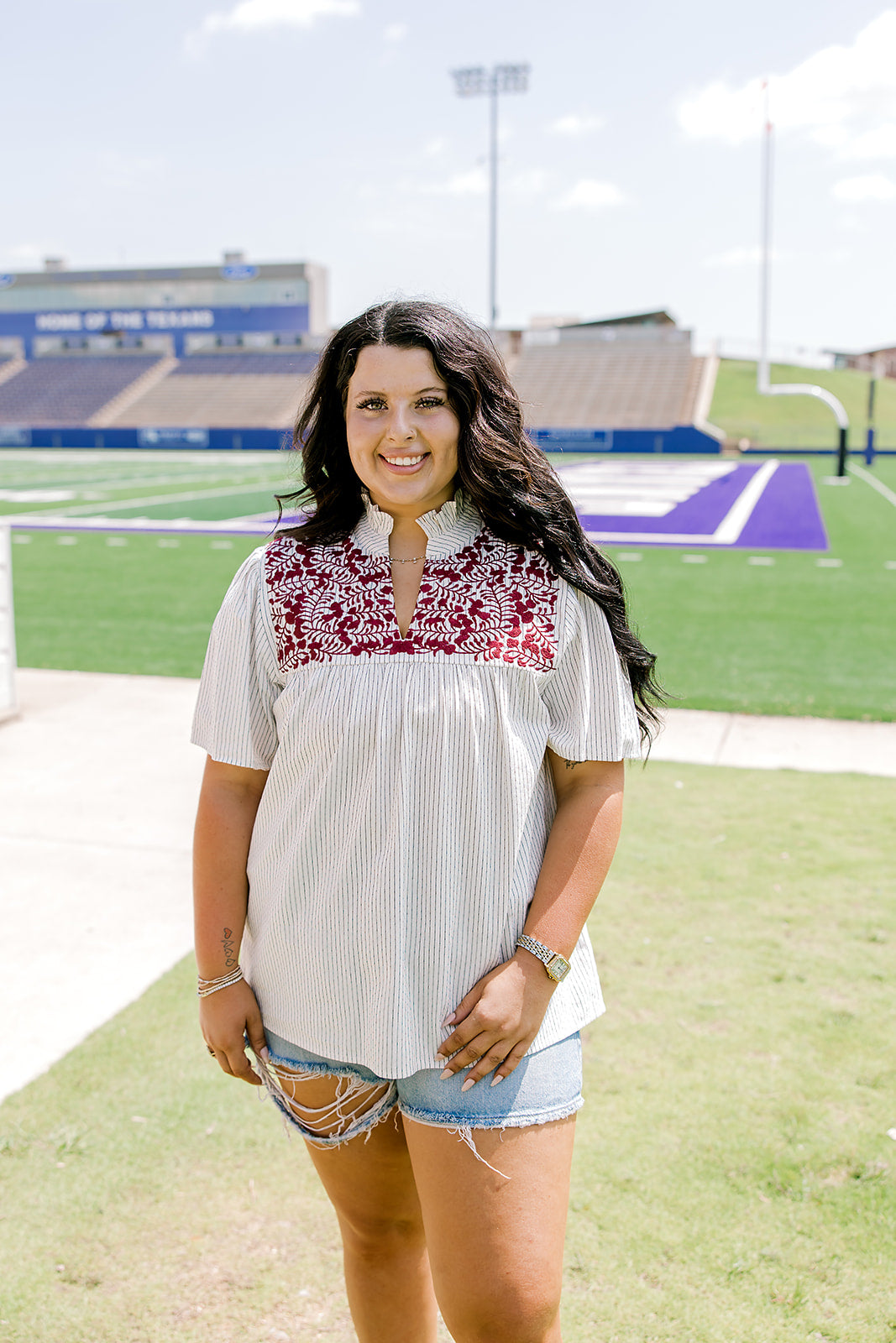 Woman with dark hair standing in a front profile position wearing the Reece Top in a lifestyle scene. The focal point of this image is the ruffled collar, v-neckline, and pleating on front of top. The model paired this top with light-wash denim shorts. 