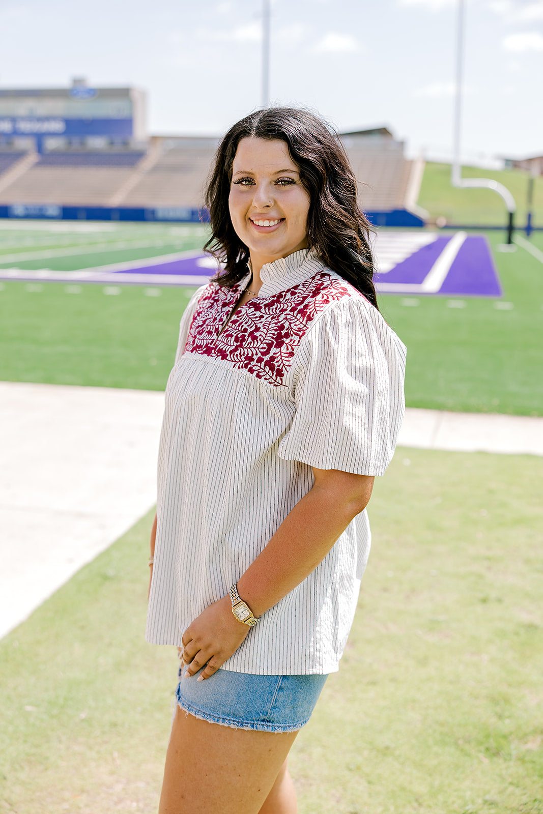 Woman with dark hair standing in a side profile position wearing the Reece Top in a lifestyle scene. The focal point of this image is the embroidery on chest as well as sleeve length and top length The model paired this top with light-wash denim shorts. 